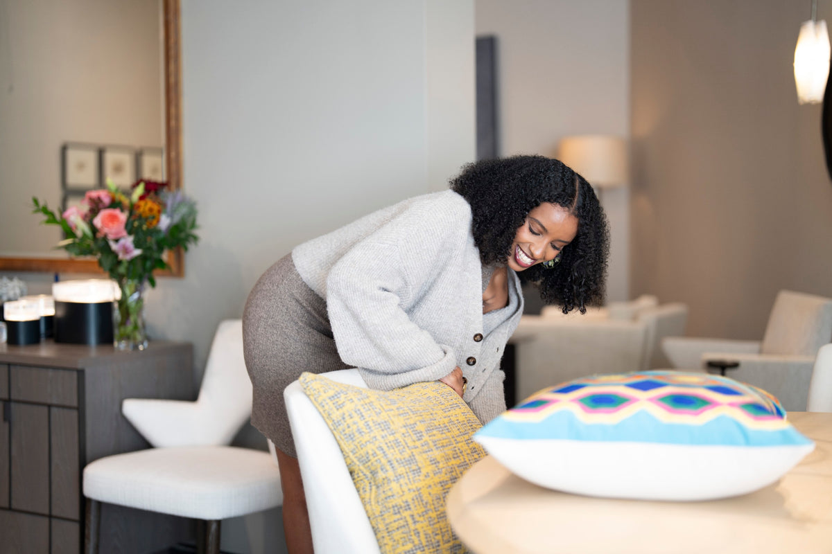 Woman setting an embroidered throw pillow on a white chair beside a vase of flowers